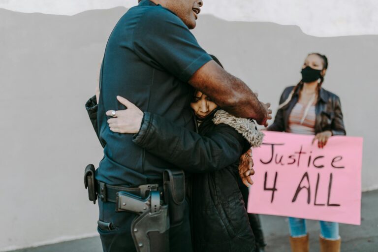 A police officer hugs a protester during a justice rally as another holds a sign reading 'Justice 4 All'.